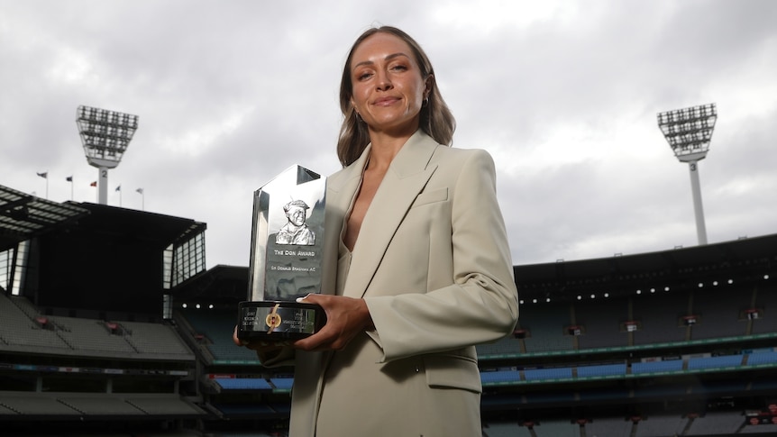 Matildas striker Kyah Simon wearing a jacket, holding a silver trophy with the light towers of the MCg in the background. 