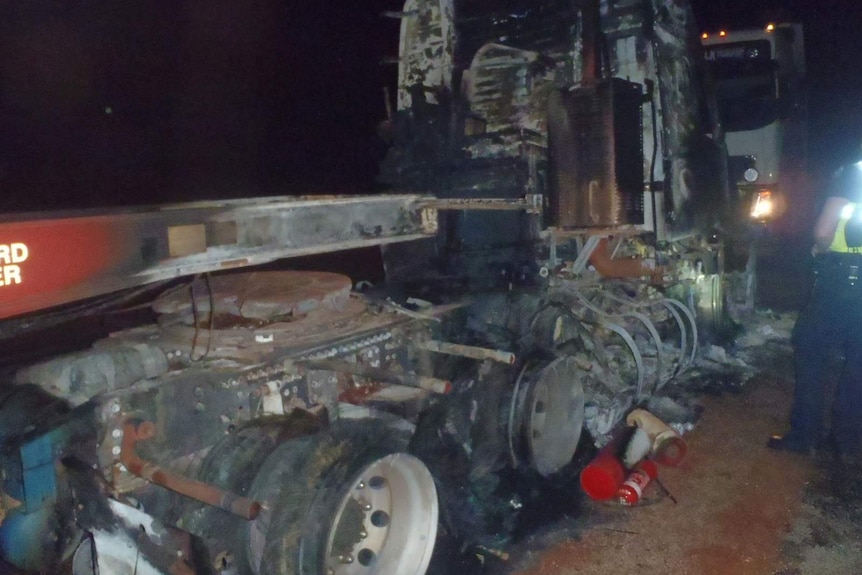 The wreckage of a road train destroyed by fire sits on the side of a highway at night with a policeman next to it.