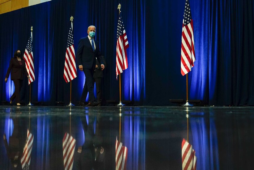 Joe Biden wears a face mask as he walks off stage in front of American flags