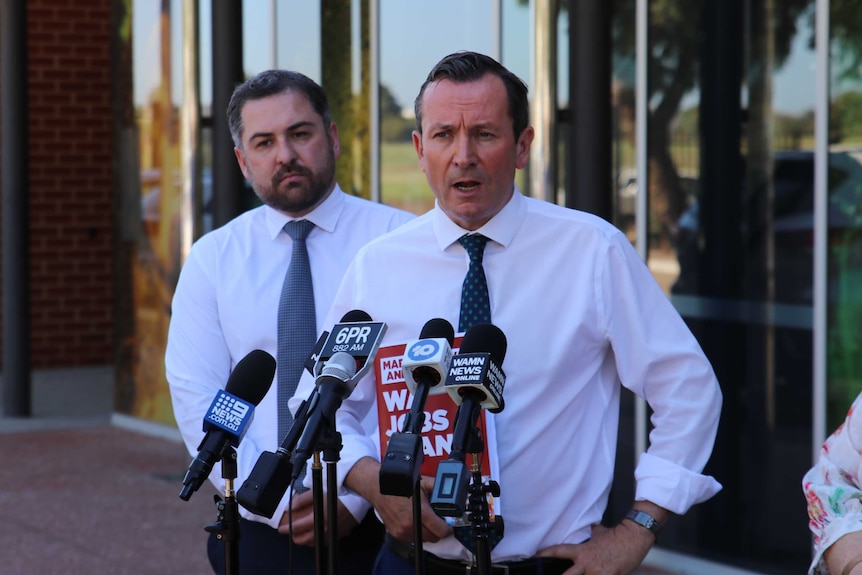 Mark McGowan holding a red election document with a man standing behind him, both standing at microphones.
