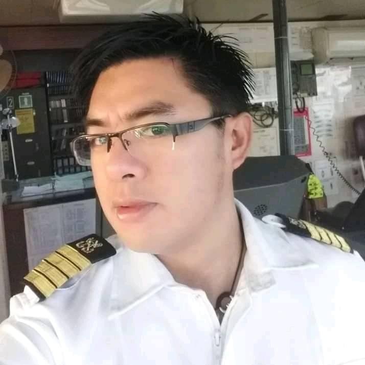 A young man with short dark hair and glasses in a captain's uniform on a ship.