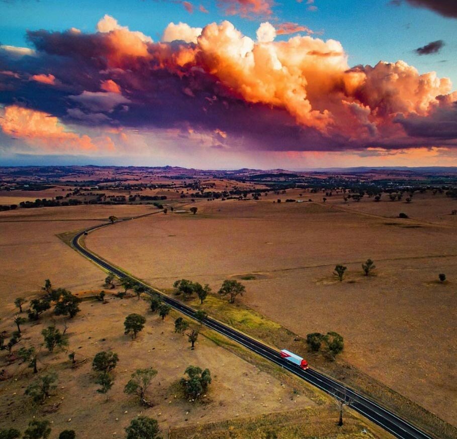 Aerial view of a semi-trailer driving along a rural highway with a township and setting sun in the distance.