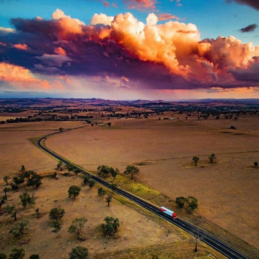 Aerial view of a semi-trailer driving along a rural highway with a township and setting sun in the distance.