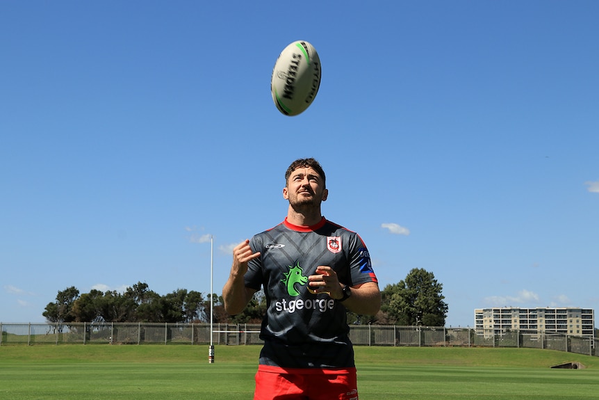 Adam Clune throws a rugby league ball in the air while standing on a football field with the posts in the background.