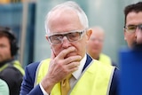 Malcolm Turnbull, wearing a yellow hi-vis vest and safety goggles over his glasses, looks downwards while covering his mouth
