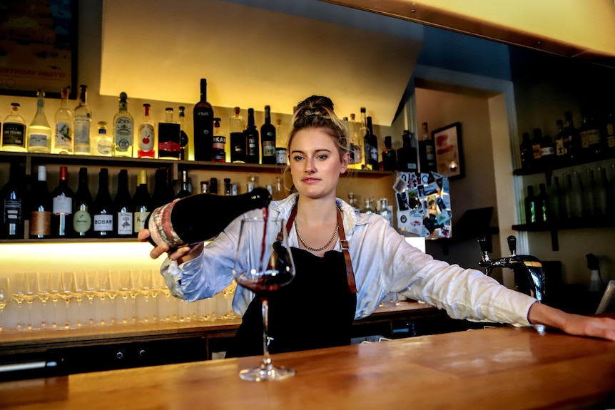 Woman with blonde hair wearing white shirt and black apron pours glass of red wine at brightly light bar
