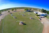 Nearly 10 helicopters sit idle on a green paddock next to a building