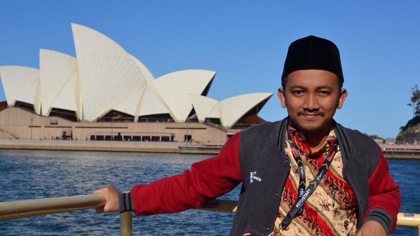A man wearing a black skullcap poses in front of the Sydney Opera House.