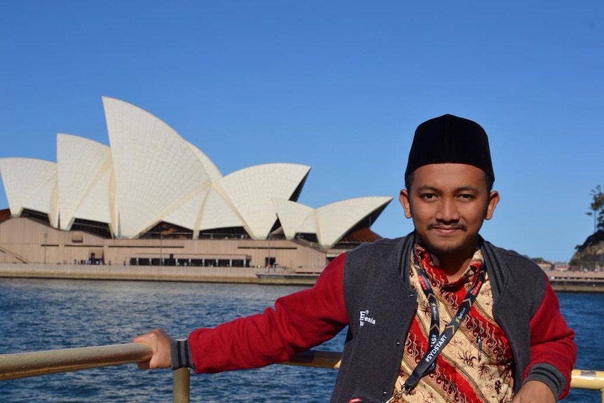 A man wearing a black skullcap poses in front of the Sydney Opera House.