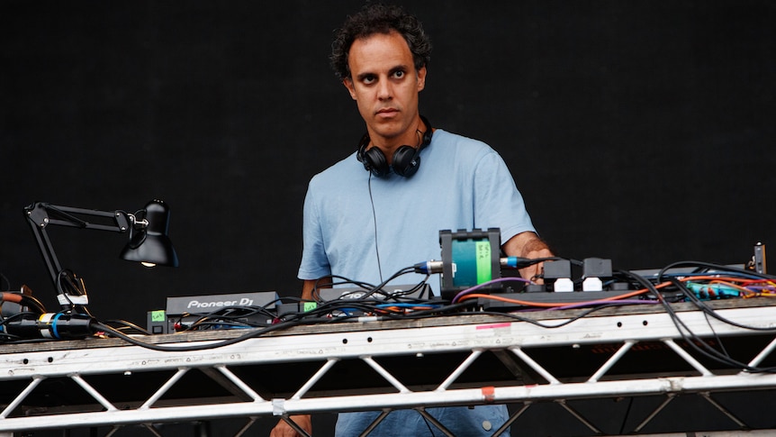 Four Tet stands behind turntables with headphones around his neck. He wears a blue shirt.