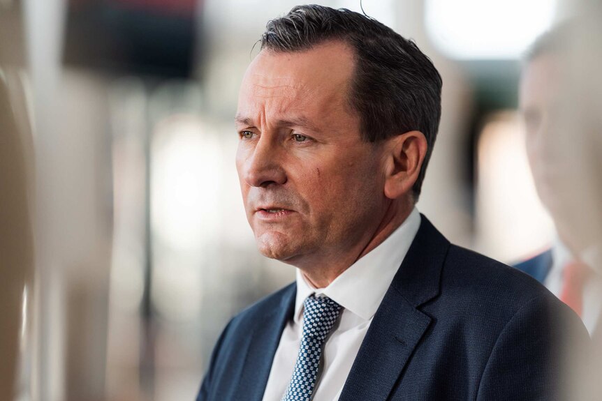 Mr McGowan speaks in front of a blurred out background, a man stands behind him.