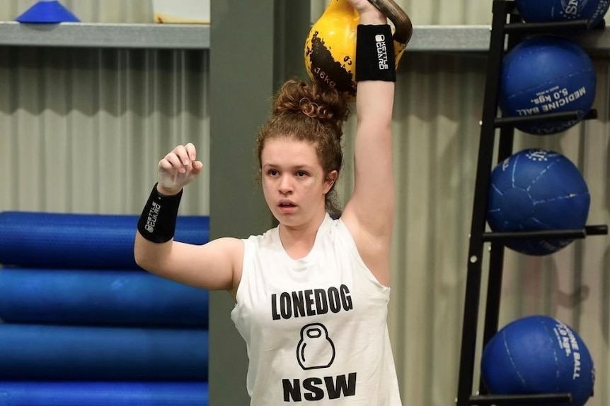 A teenage girl holds a kettlebell behind her head.