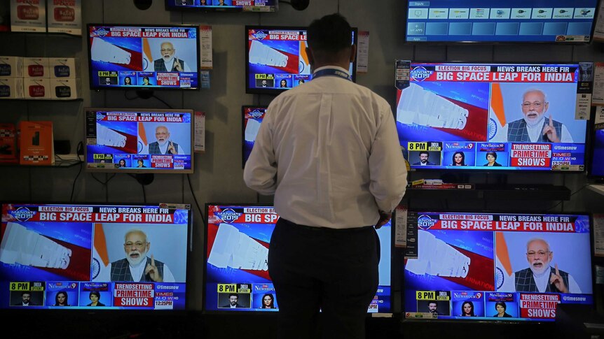 A man standing in front of multiple television screens tuned into a news report featuring Narendra Modi.
