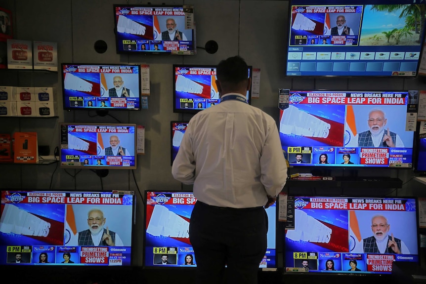 A man standing in front of multiple television screens tuned into a news report featuring Narendra Modi.