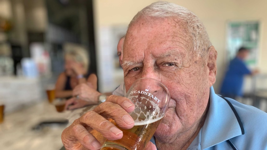 A man drinks a beer with other drinkers at tables behind him.