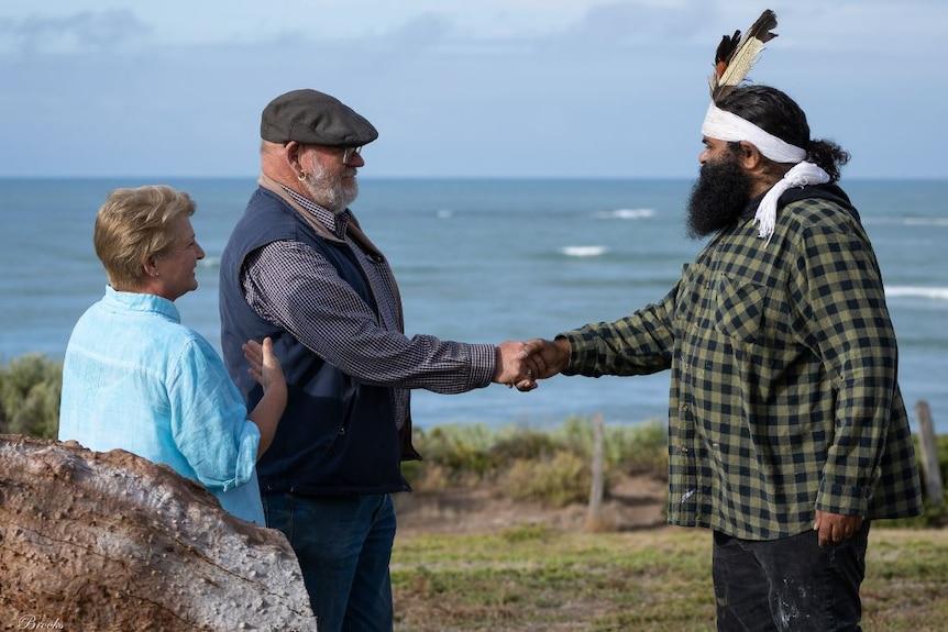 An Aboriginal elder shakes the hand of another man while a woman looks on. All stand on foreshore, sea in front.