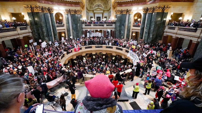 Protesters crowd a circular hall which is intricately carved in marble. 