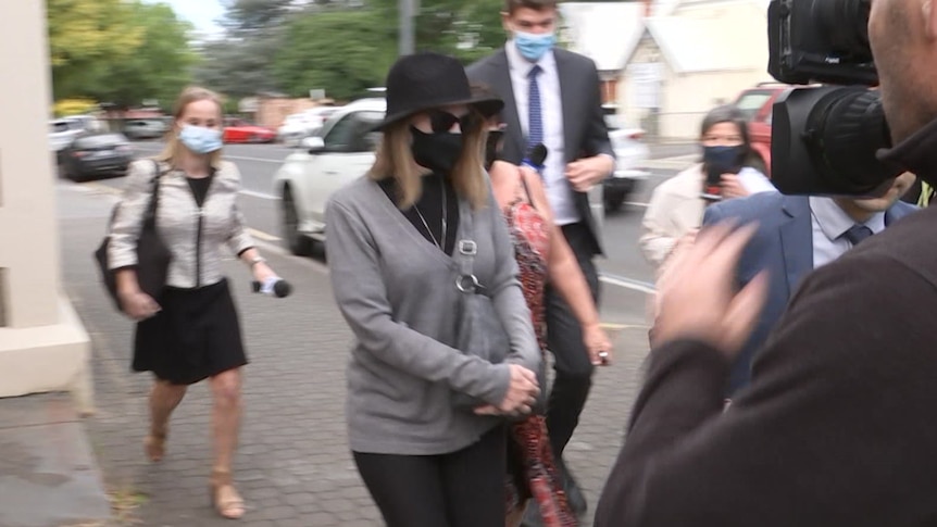 A woman wearing a black hat, face mask and sunglasses is followed by reporters