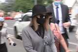 A woman wearing a black hat, face mask and sunglasses is followed by reporters