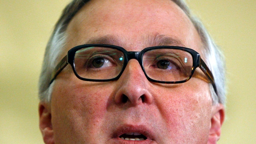 A close up headshot shows a man wearing glasses giving a speech.