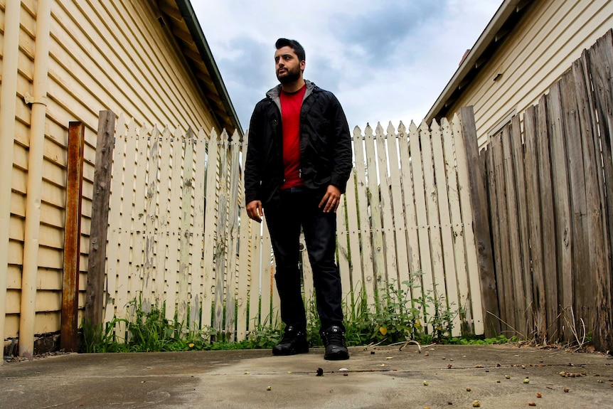 A man wearing a red shirt and black jacket stands in front of a fence with paint peeling from it.