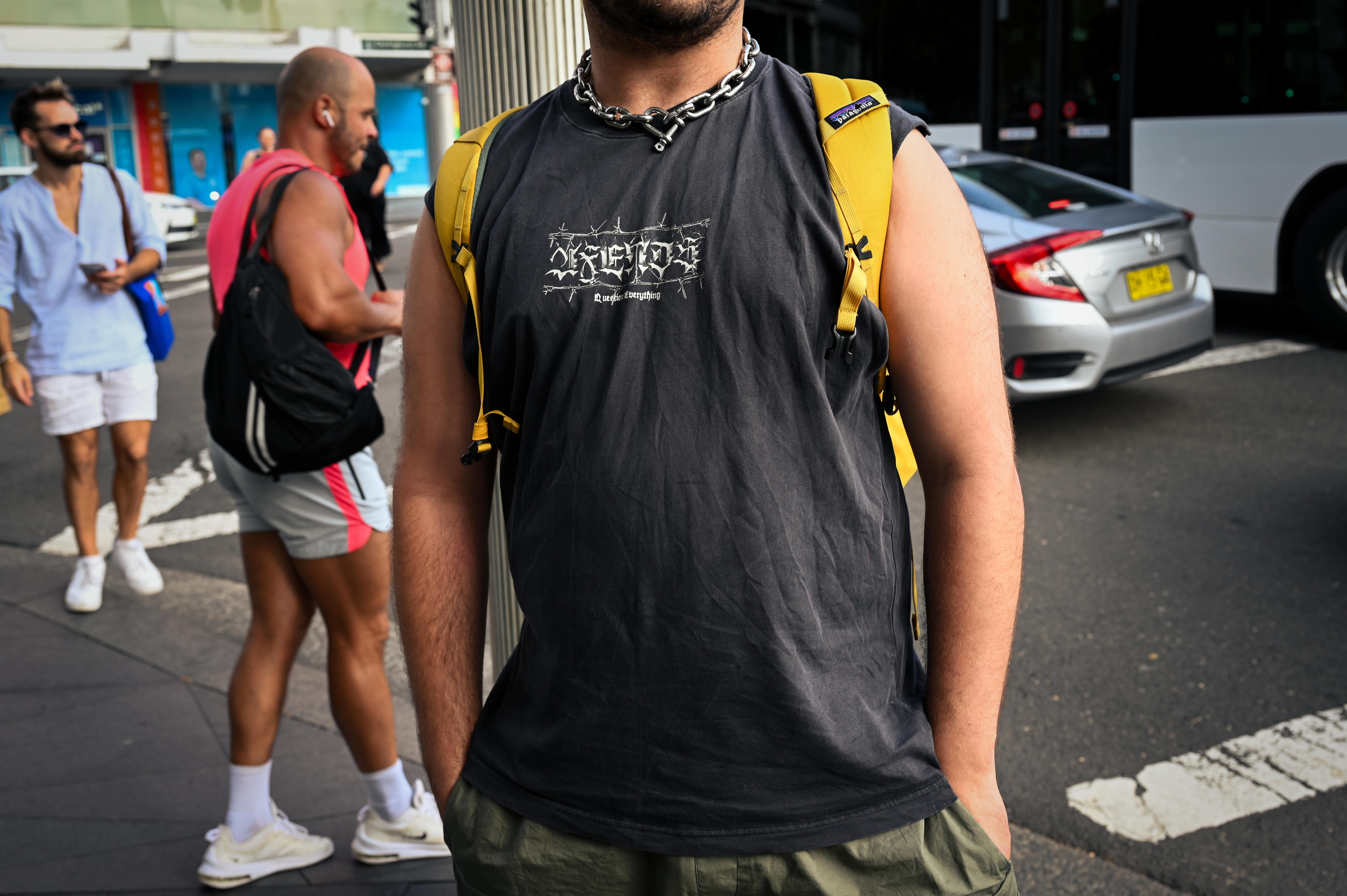 Fair-skinned man in black muscle singlet wearing a yellow backpack. He has a chain around his neck. 