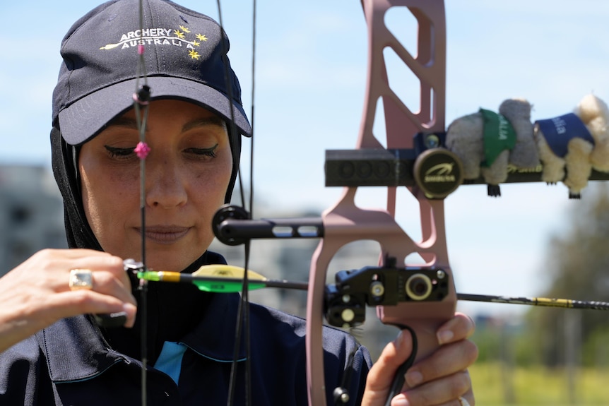 A woman wearing an Archery Australia cap looks down at her arrow as she notches it on her bow.