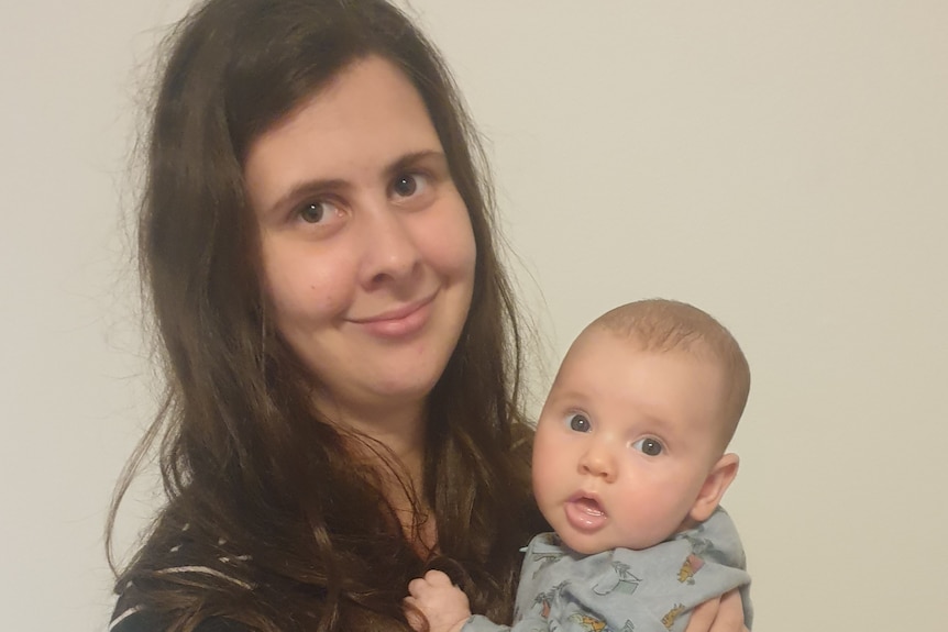 A woman holds a young baby indoors and smiles for a photo.