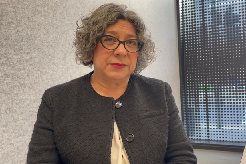 Catriona Lowe wearing a black buttoned short-coat and black glasses, sitting in front of a window.