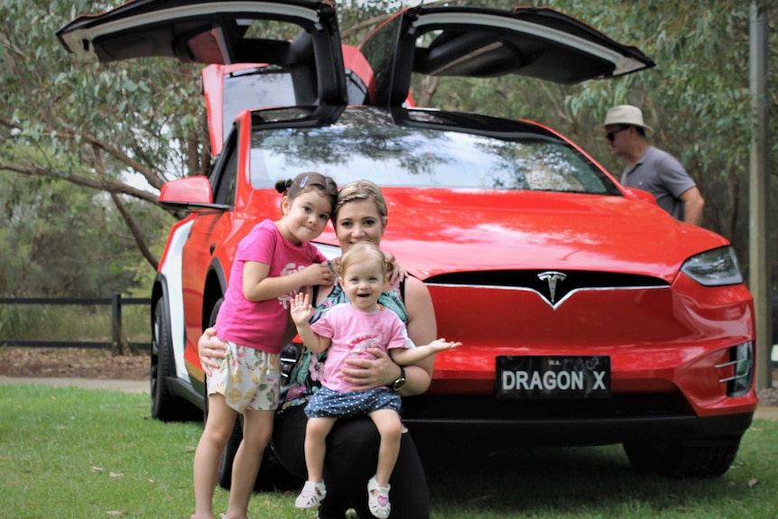 woman and two young children crouched in front of red car with sides open