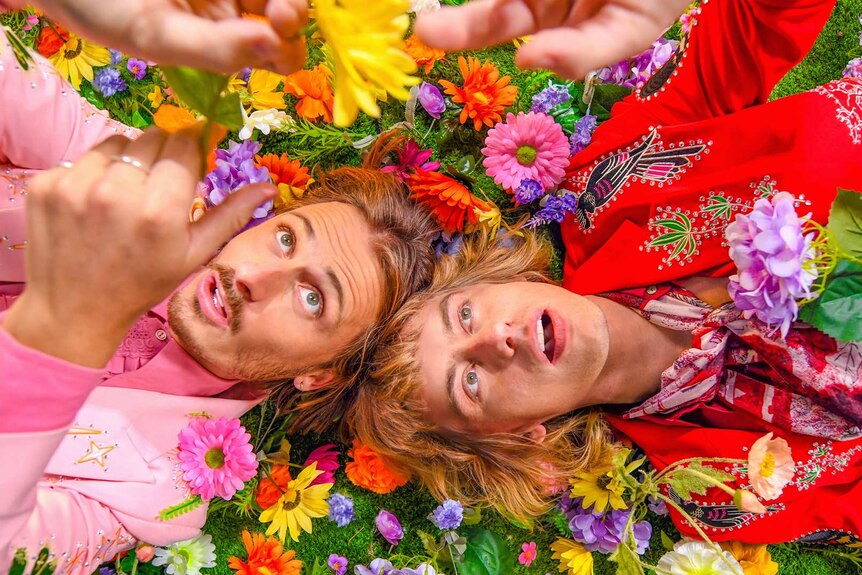 Two young men, wearing brightly coloured suits, lie on a bed of flowers.