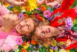 Two young men, wearing brightly coloured suits, lie on a bed of flowers.