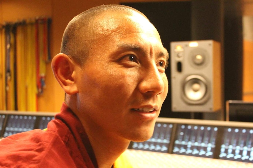 A bald monk dressed in red robes sitting in a recording studio