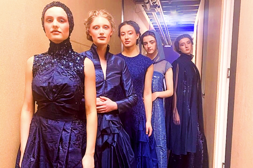 Five models stand backstage in dresses in varying shades of blue. 