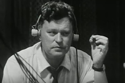 Black and white photo of man with headphones on smoking a cigar in TV control room.