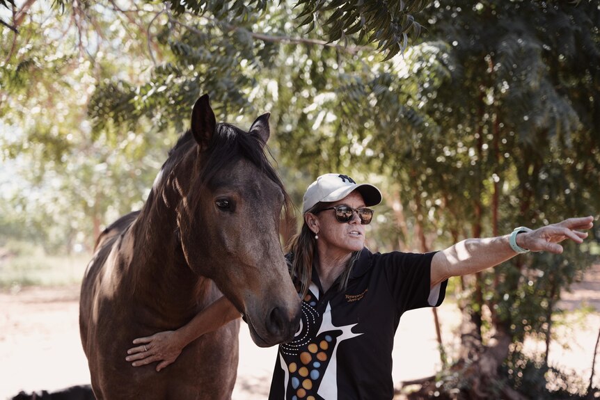 A lady wearing a hat and sunglasses standing beside a brown horse. 