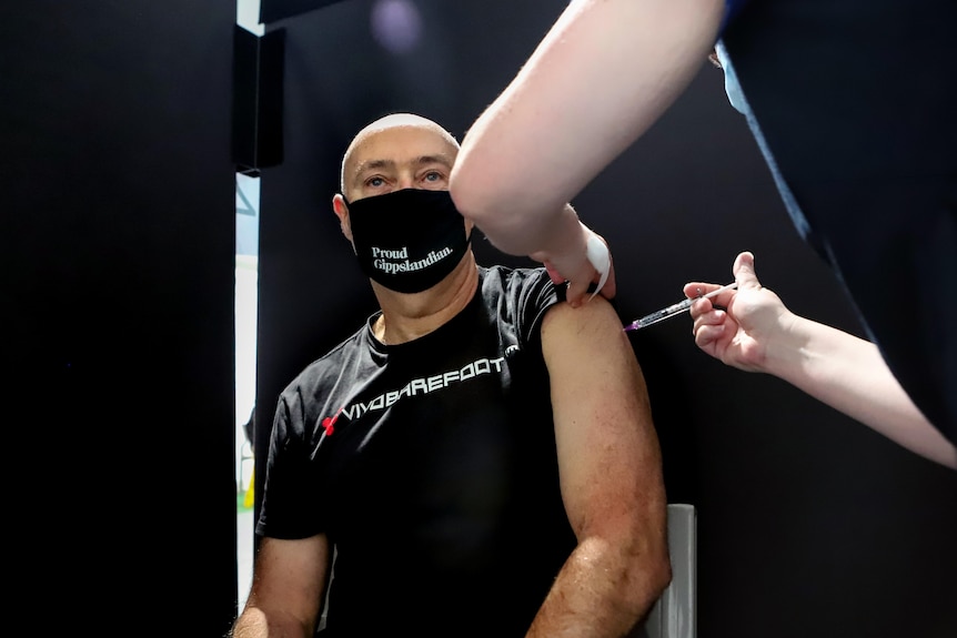 Man wearing black t-shirt and mask looks ahead as he receives injection in upper left arm from nurse