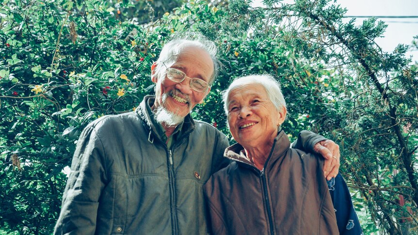 An older couple hug and smile at the camera in front of a tree