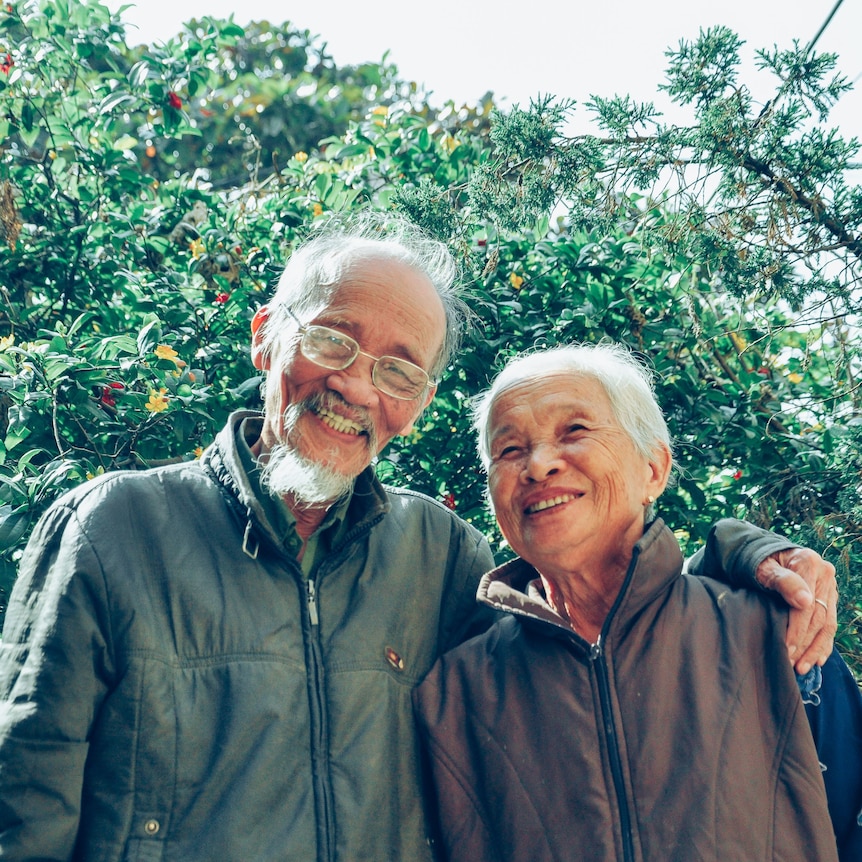 An older couple hug and smile at the camera in front of a tree