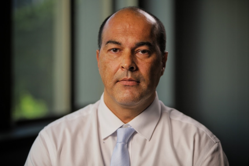 A man in a business shirt and tie is pictured in an office with a serious expression.