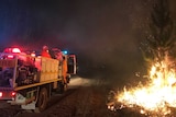 A fire truck with firefighters on board sits on a dirt road alongside burning scrub and vegetation at night.