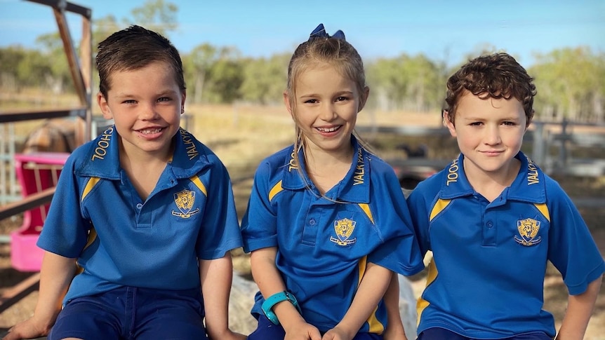 Two boys and a girl sitting on a fence in school uniforms, smiling