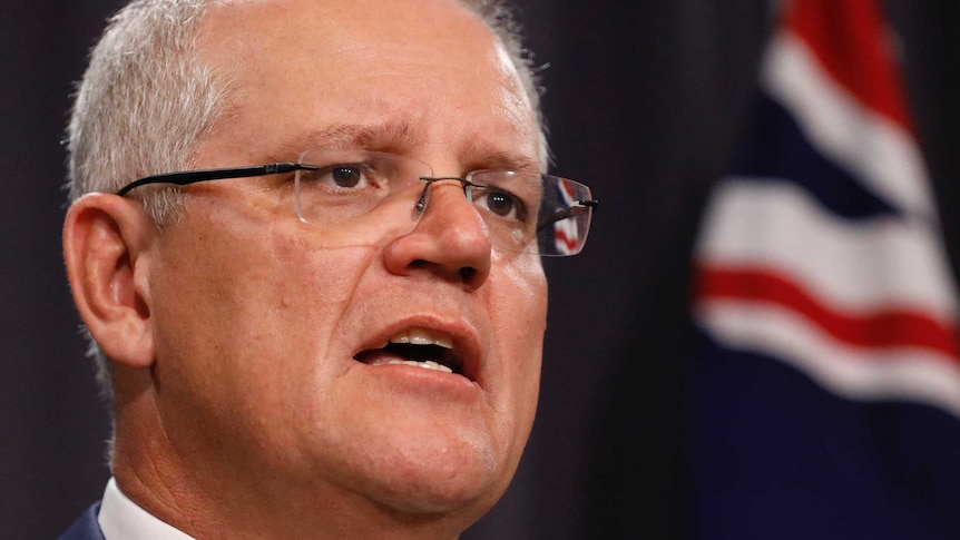 Scott Morrison stands in front of a blue curtain with an Australian flag behind him