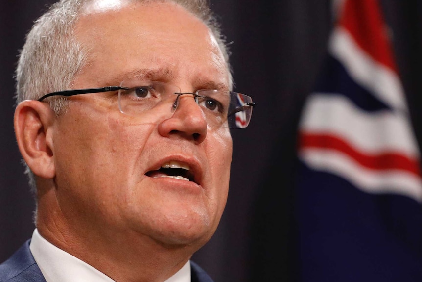 Scott Morrison stands in front of a blue curtain with an Australian flag behind him