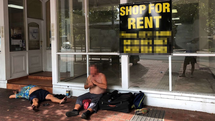 A homeless man sits and another lays sprawled on his back while camping out the front of a shop for lease.