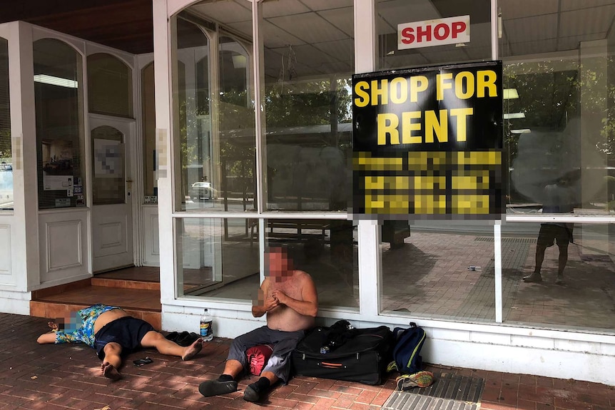 A homeless man sits and another lays sprawled on his back while camping out the front of a shop for lease.