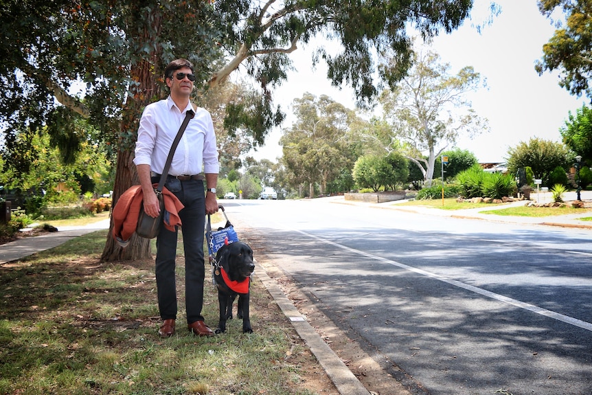 A white man with short brown hair standing on the side of a road with a black guide dog
