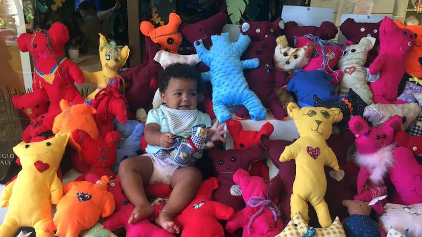 A baby lay with dozens of handmade, brightly coloured soft teddy bears.