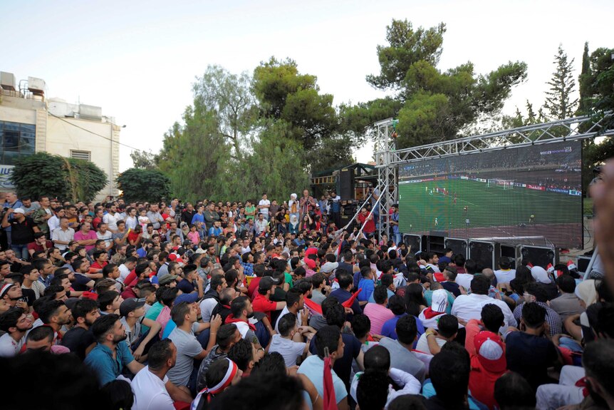 A crowd cheers as it watches a soccer game on a large TV outdoors.