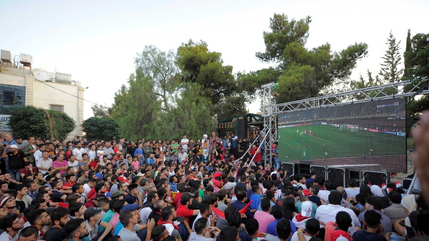 A crowd cheers as it watches a soccer game on a large TV outdoors.